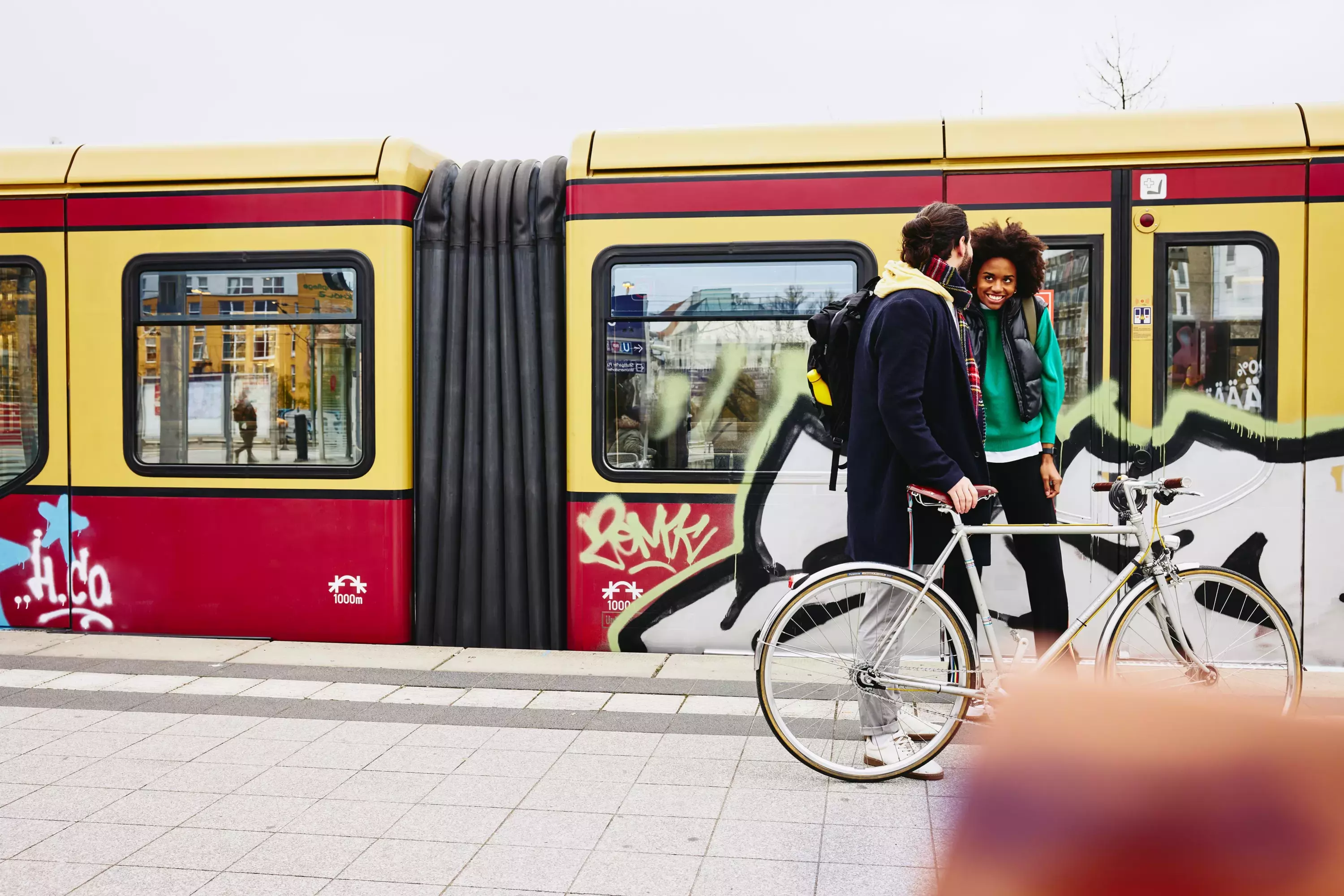 male and female standing on a train platform with a bike. Train in the background.
